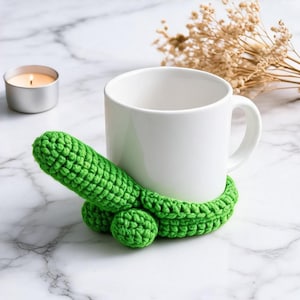 Può includere: Una tazza in ceramica bianca avvolta in una copertura all'uncinetto verde brillante. La copertura presenta una protuberanza a forma di fallo che si estende dalla base, realizzata con un punto lavorato a maglia. La tazza è liscia e cilindrica.
