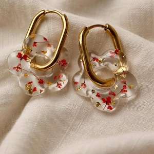 Peut inclure: Une paire de boucles d'oreilles créoles dorées. Chaque boucle d'oreille présente un charm transparent en forme de fleur avec des accents floraux rouges et des paillettes dorées. Les boucles d'oreilles sont présentées sur une surface blanche texturée.