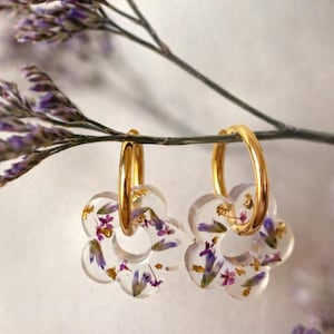 Puede incluir: Pendientes de aro dorados con colgantes de resina transparente en forma de flor. Los colgantes están rellenos de flores secas moradas y doradas. Los pendientes cuelgan de una rama con flores moradas.
