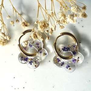 Pode incluir: Brincos de argola dourados com um design de resina transparente em forma de flor. A resina contém pequenas flores roxas e brancas, juntamente com detalhes em folha de ouro. Flores secas no fundo.