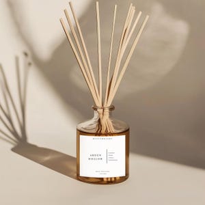 Puede incluir: Un difusor de aromaterapia de vidrio transparente con varillas de madera. La botella contiene un líquido marrón y una etiqueta blanca con las palabras "Arden Hollow" y notas de fragancia. El difusor está sobre un fondo neutro con sombras.