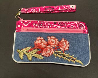 May include: A denim clutch bag with floral embroidery, a pink zip, and a matching wrist strap. The clutch has a white trim and a pink paisley patterned interior.