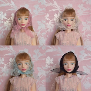May include: Four dolls with blonde hair and blue eyes, each wearing a different colored head covering. The head coverings are pink, beige, blue, and black. The dolls are wearing pink sequined dresses. The background is a pink floral pattern.
