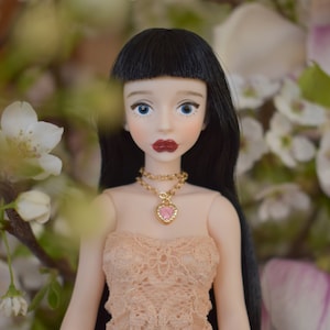 Op de afbeelding: Pop met zwart haar en blauwe ogen, gekleed in een perzikkleurige kanten jurk en een gouden ketting met een roze hartvormige hanger. De pop heeft rode lippenstift, tegen een achtergrond van witte bloemen.