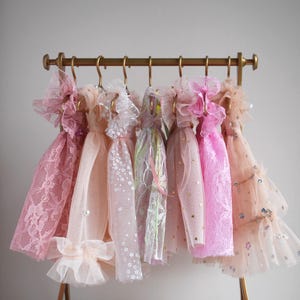 May include: A selection of miniature dresses in shades of pink and peach, displayed on a gold-coloured rack. The dresses are adorned with lace, tulle, and decorative elements, giving a delicate and fanciful appearance. The rack is approximately 30 cm wide.