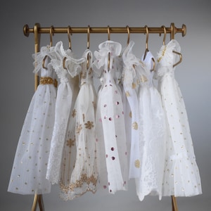 Può includere: Una collezione di abiti bianchi con dettagli dorati appesi a un appendiabiti dorato. Gli abiti presentano vari design, tra cui pizzo, tulle e abbellimenti di paillettes. L'appendiabiti ha un design semplice ed elegante.