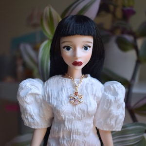 Op de afbeelding: Een pop met zwart haar en blauwe ogen, gekleed in een witte jurk met pofmouwen. De pop heeft rode lippenstift en een gouden ketting met een kleurrijke hanger. De achtergrond is wazig met groen en wit gebladerte.