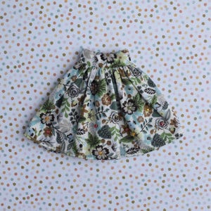 May include: A floral print skirt with a flared silhouette. The skirt features a pattern of flowers, leaves, and small embellishments in shades of green, brown, and blue on a cream background. The skirt is designed for a child.