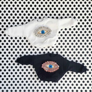 May include: Two baby sweaters, one white and one black, each decorated with a large, bejeweled eye design. The eye has a blue iris and is surrounded by sparkling crystals. The sweaters are displayed on a black and white polka dot background.