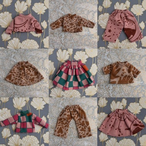 Op de afbeelding: Een verzameling kinderkleding, waaronder tops, rokken en broeken. De kledingstukken hebben verschillende patronen, zoals harten, stippen en ruiten, in roze, bruin en groen. De kleding is geplaatst op een bloemmotief achtergrond.