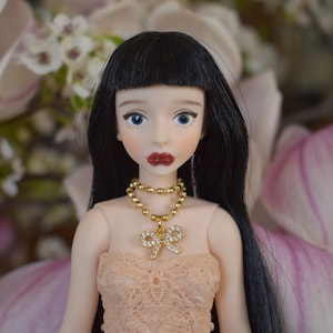 Op de afbeelding: Pop met zwart haar en blauwe ogen, met een gouden ketting met een strikvormige hanger. De pop draagt een perzikkleurige kanten jurk en rode lippenstift. De achtergrond toont zachte roze en witte bloemen.