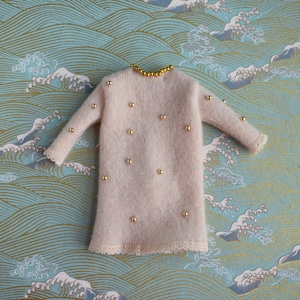 May include: A light pink, long-sleeved dress with gold bead embellishments and a lace trim. The dress has a round neckline with gold beads and is decorated with evenly spaced gold beads. The background features a blue and white wave pattern.