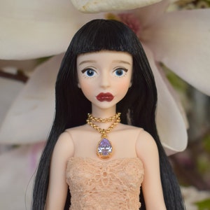 Op de afbeelding: Een pop met zwart haar, blauwe ogen en rode lippenstift. De pop draagt een gouden ketting met een paarse druppelvormige hanger en een perzikkleurige kanten jurk. De achtergrond is een wazige witte bloem.