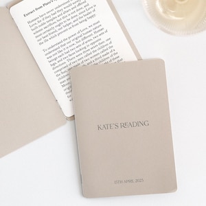 Könnte beinhalten: Ein beiges Büchlein mit dem Text "KATE'S READING" und dem Datum "25. April 2023" ist neben einem Weinglas geöffnet. Das Büchlein ist auf einer Seite mit gedrucktem Text geöffnet. Der Hintergrund ist weiß.
