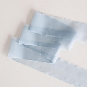 May include: A light blue ribbon with frayed edges is arranged on a white surface. The ribbon is folded over itself in a layered pattern, with the ends extending to the right. The ribbon's color is a soft, pastel blue.