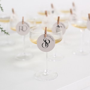 May include: Several champagne glasses filled with a golden liquid, each with a round tag attached by a small wooden clip. The tags display initials and the date 21.06.2027, indicating a special occasion or celebration.