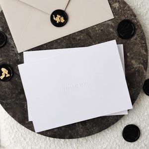 May include: A stationery set featuring a beige envelope, white cards with "THANK YOU" embossed text, and black wax seals with gold accents. The items are arranged on a dark, textured surface.