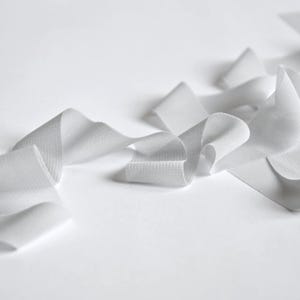 May include: Close-up of white ribbon arranged in a flowing, abstract pattern against a white background. The ribbon has a textured appearance, with soft folds and curves. The image has a minimalist aesthetic.