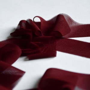 Op de afbeelding: Close-up van een diep bordeauxrood lint, mogelijk fluweel of een vergelijkbare stof, met een licht gestructureerd oppervlak. Het lint is in losse plooien en bochten gerangschikt, waardoor een gevoel van beweging ontstaat tegen een witte achtergrond. Ideaal voor knutselen of decoratie.
