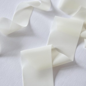 Op de afbeelding: Close-up van ivoorkleurig lint, gedrapeerd over een witte ondergrond. Het lint heeft een gladde, doorschijnende uitstraling met zachte plooien en bochten. De afbeelding benadrukt de delicate textuur van het lint.