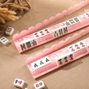 May include: Two long, scalloped-edge pink marble-effect acrylic racks holding white Mahjong tiles with red, green, and black characters. Several loose Mahjong tiles are scattered nearby, displaying various numerical and symbolic markings in red and green.