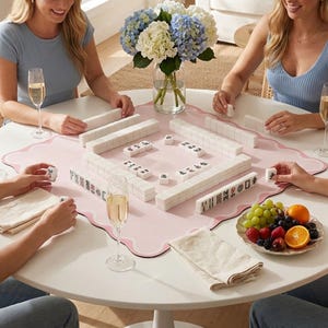 May include: A square, light pink mahjong mat with a scalloped edge, featuring a set of white mahjong tiles arranged in rows and stacks on the surface. The mat is placed on a round white table alongside glasses of champagne, napkins, and fruit.