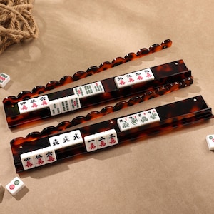 Può includere: Due lunghi supporti rettangolari per tessere del mahjong con un motivo a tartaruga marrone scuro e bordi superiori smerlati. Ogni supporto contiene una fila di tessere bianche con caratteri e simboli cinesi neri e rossi. Tessere sparse circondano i supporti.