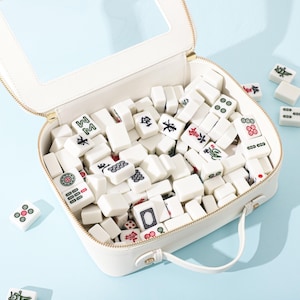 Könnte beinhalten: Ein weißer Tragekoffer mit Reißverschluss und transparentem Sichtfenster im Deckel, gefüllt mit einer Sammlung weißer, rechteckiger Mahjong-Spielsteine mit verschiedenen eingravierten schwarzen, roten und grünen Symbolen und Schriftzeichen.