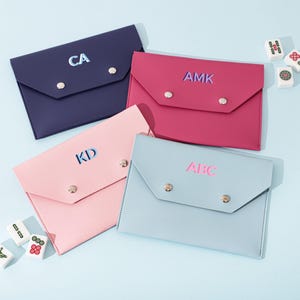 May include: Four rectangular envelope-style pouches in navy, magenta, light pink, and light blue. Each pouch features two silver-tone snap closures and personalized lettering: CA, AMK, KD, and ABC. Small white mahjong tiles are scattered nearby.