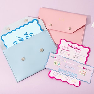 Pode incluir: Uma bolsa azul-clara estilo envelope com fecho de pressão contendo um cartão recortado, uma bolsa rosa estilo envelope e dois cartões de pontuação de Mahjong recortados com texto incluindo "MAHJONG" e "Laura", junto a três peças brancas de Mahjong.