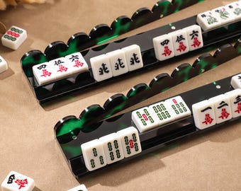 Könnte beinhalten: Zwei lange Mahjong-Fliesenhalter aus schwarzem Acryl mit einem gewellten, durchscheinend grünen Rand. Jeder Halter enthält eine Reihe weißer Mahjong-Steine mit schwarzen und roten chinesischen Schriftzeichen sowie grünen Bambussymbolen.