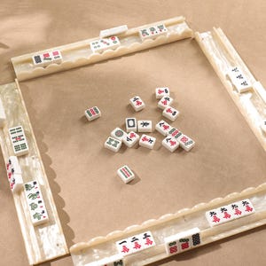 May include: A Mahjong game set featuring four off-white, pearlescent tile racks with scalloped edges and a collection of rectangular tiles. The tiles are white with various engraved symbols, characters, and numbers in red, green, and black ink, scattered across the center and arranged on the racks.