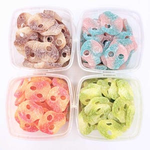 Könnte beinhalten: Vier transparente Plastikbehälter gefüllt mit bunten, schädelformigen Süßigkeiten. Die Süßigkeiten sind in Braun-, Rosa- und Blautönen, Orange und Rosa sowie Grün gehalten. Jedes Bonbon ist mit Zucker überzogen.