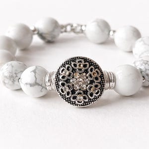 May include: White marble bead bracelet with a decorative silver-tone centerpiece. The centerpiece features a floral design with clear crystal accents. The bracelet is strung on a silver chain.