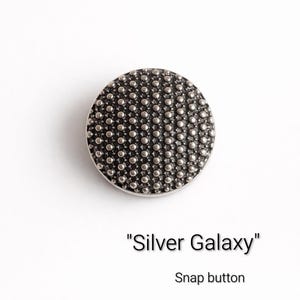 Puede incluir: Un botón de presión redondo plateado con una superficie texturizada, con un patrón de pequeños puntos en relieve. El botón está etiquetado como "Silver Galaxy" y está diseñado para sujetar prendas o accesorios.