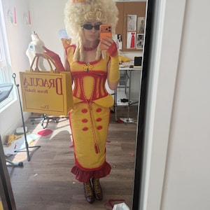 Könnte beinhalten: Ein gelbes und rotes Kostüm mit passender Perücke und Hut. Das Kostüm hat ein korsettartiges Oberteil und einen Rock mit rotem Besatz. Die Person hält eine gelbe Tragetasche mit dem Text "Dracula".