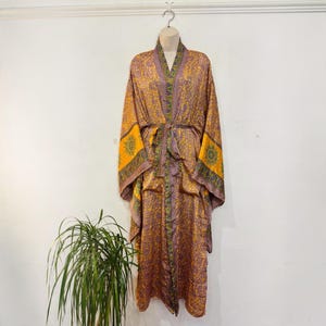 May include: A long-sleeved kimono robe featuring a vibrant pattern of yellow and purple floral motifs. The garment includes a matching fabric tie belt at the waist and a contrasting border along the front opening and sleeve cuffs.