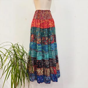 Op de afbeelding: Een enkellange, gelaagde maxirok met een gesmokte elastische tailleband en een levendig patchworkontwerp. De rok toont diverse kleurrijke paisley- en bloemenpatronen in tinten rood, blauwgroen, blauw, zwart en crème over de gehele stof.