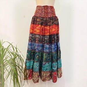 Op de afbeelding: Een lange, gelaagde maxirok met een gesmokte elastische tailleband en een levendige, meerkleurige paisleyprint. De rok is opgebouwd uit horizontale stroken stof in tinten rood, oranje, blauw, groenblauw en bruin, wat zorgt voor een vol silhouet.