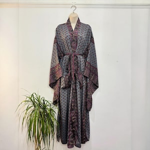 May include: A long-sleeved, floor-length robe in a muted grey tone, featuring a repeating small floral pattern throughout. The garment includes a wide, dark purple paisley-patterned border along the front opening, cuffs, and a matching tie belt.