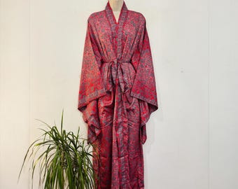 May include: A long-sleeved robe featuring a vibrant red and grey intricate paisley pattern. The garment includes a matching fabric tie belt at the waist, wide sleeves, and a shawl collar, displayed on a mannequin against a plain white wall.