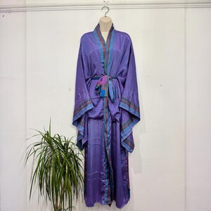 Könnte beinhalten: Ein bodenlanger violetter Kimono-Bademantel mit einem sich wiederholenden geometrischen Muster in Dunkelblau und kleinen roten Akzenten. Das Kleidungsstück verfügt über weite Ärmel, einen passenden Bindegürtel in der Taille und kontrastierende Säume.