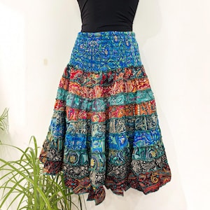 May include: A tiered midi skirt featuring a smocked elastic waistband and a vibrant patchwork design. The skirt displays a variety of colourful paisley and floral patterns in shades of blue, red, orange, green, and black, with ruffled seams.