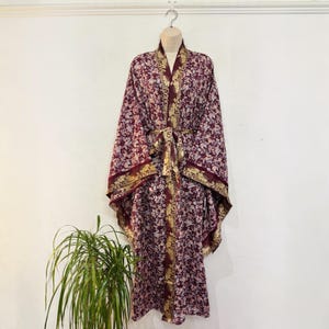 May include: A floor-length kimono robe featuring a deep burgundy base with a dense floral pattern in white and light blue. The garment includes a wide, contrasting gold-toned border along the front opening and sleeves, with a matching tie belt.