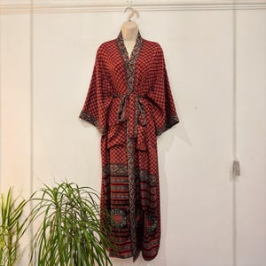May include: A long, red patterned robe with a matching belt. The robe features a geometric design in black and red, with decorative borders along the edges and sleeves. The robe is displayed on a mannequin against a white wall.