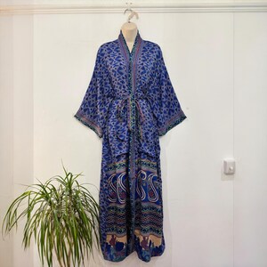 May include: A long, blue patterned robe with a matching belt. The robe features a geometric design on the upper portion and a paisley pattern on the lower portion. The robe is hanging on a hanger against a white wall.
