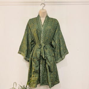 May include: A knee-length kimono robe featuring a dark green and gold paisley pattern. The garment has wide, three-quarter length sleeves, a V-neckline, and a matching fabric tie belt at the waist. The fabric displays a repeating intricate design.