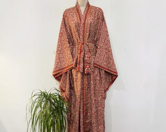 May include: A floor-length kimono robe featuring a repeating intricate pattern in shades of red, orange, and cream. The garment has wide sleeves, a front tie closure, and a structured collar, displayed on a mannequin against a plain wall.