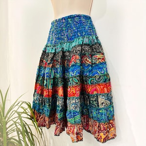 May include: A tiered midi skirt featuring a smocked elastic waistband and a vibrant patchwork design. The skirt is composed of horizontal fabric panels in various colors including blue, red, orange, and black, decorated with intricate paisley patterns.