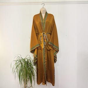 Op de afbeelding: Een mosterdgele kimono-badjas met lange mouwen en een herhalend geometrisch patroon in donkerbruin. Het kledingstuk is voorzien van een bijpassende stoffen ceintuur in de taille en een decoratieve blauwgroene en witte rand langs de voorzijde.